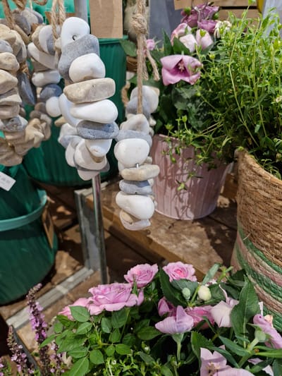 The image shows a display of decorative items and plants, likely in an outdoor market or garden center. In the foreground, a strand of polished stones, strung together with twine, hangs vertically. The stones vary in size, shape, and color, ranging from white and grey to beige. Behind this, several potted plants are visible. To the right, a woven basket holds a bushy green plant with small leaves and budding flowers. To its left, a light pink, metallic pot contains another plant with delicate pink flowers and green foliage. In the background, more greenery and hints of purple flowers are visible, as well as large green planters. The scene is illuminated by natural light, casting shadows on a wooden surface, which appears to be a tabletop or display stand. There are no people visible in the image. The overall impression is one of a botanical or artisanal retail setting.