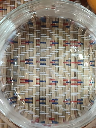 The image shows a close-up, overhead view of a woven placemat with a red, white, and blue striped pattern. The placemat is underneath a clear glass bowl or coaster, which is reflecting light and distorting the view of the woven material. The woven pattern consists of thin, light-colored strands interlaced with thinner strands of red and blue. The glass creates a slightly magnified and warped effect on the placemat's texture. There are some shiny highlights on the surface of the glass, indicating a light source. The overall impression is a still life composition, focusing on the texture and pattern of the placemat as seen through the glass. No people, specific activities, time of day, weather, or location cues are visible. There is no visible text in the image.