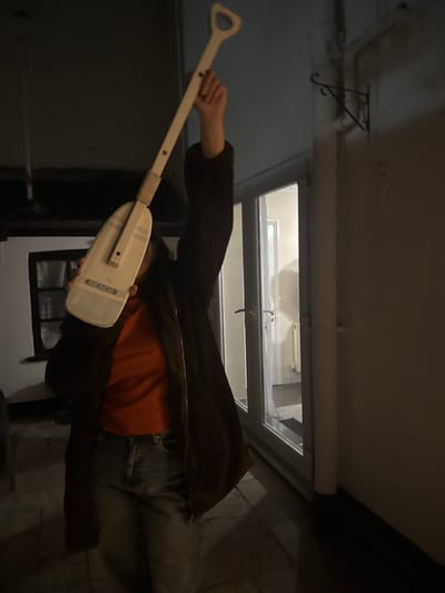 A person is lying on their side, holding a white paddle in their left hand. The paddle has a "BEACH" label near the bottom. The person is wearing a brown jacket over an orange shirt and grey jeans. The setting is indoors, with a skylight above. Through the skylight, a snowy landscape is visible. A metal bracket is fixed to the ceiling. The location cannot be determined as Gorleston-on-Sea, United Kingdom.