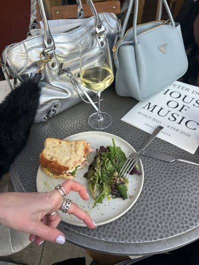 The scene depicts a casual outdoor dining setting in the City of Westminster, United Kingdom, specifically at or near Serpentine South, as indicated by printed material. A textured dark grey metallic table holds a white plate containing a half-eaten sandwich with a light-colored filling and a mixed green salad with seeds, speared by a silver fork. A knife rests beside the plate. To the right of the plate, a wine glass contains a clear pale yellow liquid. Two handbags are prominently displayed: a crinkled silver bag with multiple zippers and buckles on the left, adorned with a black fluffy charm, and a light blue Prada shoulder bag with a gold logo on the right. A white brochure titled "PETER DO HOUSE OF MUSIC" with dates "10 OCT 2025 – 8 FEB 2026" and "SERPENTINE SOUTH" is visible behind the light blue bag. A person's left hand, with light purple nail polish and multiple silver rings on the index, middle, and ring fingers, is positioned in the foreground, with the index and middle fingers slightly separated. A dark, possibly fur-trimmed, sleeve is visible on the far left, belonging to the same individual.