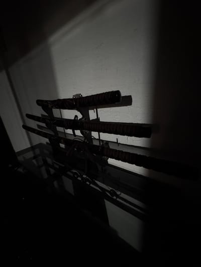 The media file displays a dark, indoor scene featuring a collection of swords, likely katanas, mounted vertically on a wall rack. Approximately five to six swords are discernible, arranged in a row. Each sword features a dark hilt wrapped tightly with a distinctive red or deep crimson thread, and black scabbards. Faintly visible on the lower section of some scabbards are intricate, ornate designs, possibly depicting dragons or mythical figures, though details are obscured by shadow.

The swords are held by a dark, possibly metallic, wall-mounted rack that casts prominent shadows. The wall itself is a light gray or off-white hue with a visible rough texture, similar to stucco or painted concrete. A strong, directional light source, originating from the left side of the frame, illuminates the wall and the handles of the swords, creating dramatic and elongated shadows that stretch across the wall to the right. The right portion of the image is enveloped in deep shadow, rendering objects in that area almost completely black and indistinct.

The overall impression is one of a dimly lit space, suggesting either nighttime or a room with minimal ambient light. No human subjects, specific activities, or discernible text are present. The atmosphere is stark and somewhat mysterious due to the stark contrast between the illuminated and shadowed areas and the nature of the displayed weaponry. The location context provided is Cuernavaca, Mexico, but no visual elements within the image directly confirm this geographical setting.