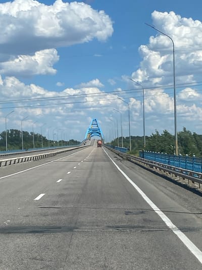 The image shows a perspective view of a highway bridge crossing a body of water, likely a river, under a bright, cloudy sky. The bridge features a distinctive blue arch structure in the distance and is lined with lampposts. The highway itself is asphalt with visible white lane markings. On the right side of the bridge, a blue metal railing is present. A truck is visible driving on the bridge in the distance. The surrounding environment appears to be a rural or semi-rural area with trees lining the banks of the waterway. There is no indication that this scene is located in Aqsu city administration, Kazakhstan.