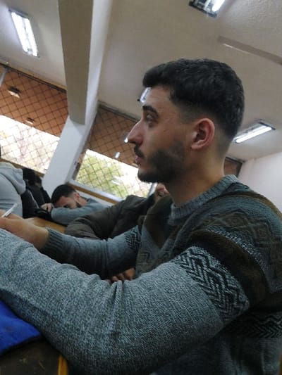 The image captures an indoor scene, likely a classroom or lecture hall, located within Ash-Shaghour Municipality, Syria. In the foreground, a young adult male is positioned in profile, facing right, with his mouth slightly open. He has short, dark hair styled with a fade and a neatly trimmed beard and mustache. He is wearing a light grey knitted sweater with horizontal bands of dark grey and brown intricate geometric patterns across the upper chest, shoulders, and outer sleeves. His right forearm rests on a dark, possibly wooden, surface, beneath which a blue fabric item is visible. His left hand, partially visible, holds a white writing implement.

In the mid-ground, behind the primary subject, another male figure is seen resting his head on a surface, dressed in a grey long-sleeved top and a dark jacket. Additional indistinct figures are present further in the background. The room's environment includes a light-colored ceiling fitted with multiple fluorescent tube lights. Light-colored walls are visible, alongside windows featuring a grid pattern and light-brown Venetian blinds. Green foliage is discernible through the windows, indicating an outdoor view.
