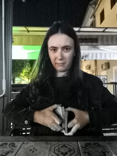 A person with long, dark hair and a dark denim jacket sits at a table, holding a deck of cards in their hands. The person's face is partially obscured by their hair, and they are looking towards the camera with a neutral expression. The background appears to be an outdoor seating area, possibly a cafe or restaurant, with a sign that reads "Pub Academia". The lighting is dim, suggesting it is nighttime or evening. The table in front of the person has a patterned surface. The overall scene is dimly lit, with a green glow visible in the background from some artificial lighting.