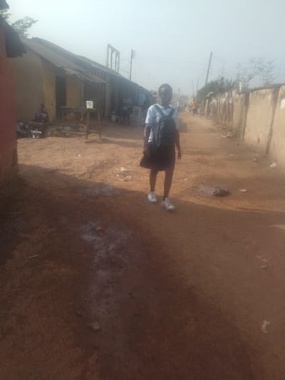 The image shows a young person, possibly a school student, walking down a dirt road in New Karu, Nigeria. The road is dusty and uneven with a few puddles of water. The student is wearing a white shirt and black skirt, and carries a black backpack. They are wearing white sneakers. There are several buildings to the left, likely residential houses, and a wall on the right. The person is walking away from the camera. The environment is moderately lit and there are no other people in the scene. The student seems to be on their way to school, as the backpack suggests they are carrying books or other school supplies. The time of day is likely morning or afternoon, based on the brightness of the scene and the presence of some shadows. There are no vehicles or other forms of transportation visible. The image depicts a common scene in many parts of New Karu, highlighting the simple lifestyle and reliance on walking as a primary mode of transportation in the area.