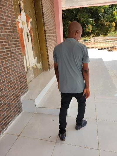 In Ebenebe, Nigeria, a brightly lit outdoor scene depicts a single male subject walking away from the camera within a tiled covered walkway. The individual has a shaved head, wears a grey short-sleeved polo shirt, black trousers, and dark blue open-toed sandals. To the left, a reddish-brown brick wall features a tall, golden-framed door, upon which is a relief or painted depiction of a robed figure, possibly a saint or Jesus, holding a staff. The walkway floor consists of large, light-colored tiles, and the roof overhead is supported by light pinkish beams. Beyond the covered area, green tree foliage is visible in the mid-ground, with a metal fence and an open, lighter-toned area forming the distant background.