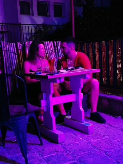 Two individuals, a woman and a man, are seated at a small, rustic wooden table in an outdoor setting illuminated by purple and pink lights. The woman, with dark hair, is wearing a dark spaghetti-strap dress and is looking towards the man. The man, with short dark hair and a beard, is wearing a red t-shirt. On the table are two glasses, one with amber liquid, a bottle, and some papers. A dark metal chair is partially visible on the left of the frame. Behind them, a bamboo fence and a building with visible windows can be seen. The ground appears to be paved with irregular stones. The scene suggests they are at a bar or restaurant. The location is identified as Herceg Novi, Montenegro, likely an outdoor seating area of a establishment in the city.