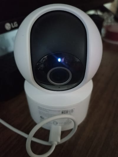 This image depicts a white, spherical-headed security camera with a black front panel, positioned on a dark brown wooden surface in the city of Serpukhov, Russia. The camera's lens is centered on the black panel, and a small, illuminated blue LED indicator is visible above it. The spherical head rests on a cylindrical white base. A white USB-A cable is plugged into the rear of the camera's base, looping towards the foreground. Text and regulatory markings, including "NOM," are partially visible on the camera's base. In the blurred background to the left, a dark electronic device displaying an "LG" logo is present. To the right, a blurred red object is partially visible. No people are present, and no actions or interactions are depicted.