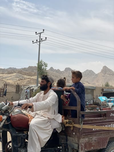 A man drives a modified motorcycle, which has a large, rusty cargo bed attached to its rear, functioning as a multi-person vehicle. He wears a light-colored traditional outfit (shalwar kameez) and a patterned skullcap, with a dark beard, and is looking straight ahead, hands on the handlebars. In the back of the open-air cargo bed, two young children are visible. A boy in a dark blue outfit stands at the edge of the bed, gripping the frame and looking forward. Behind him, another child with pigtails is partially visible.

The scene unfolds under a cloudy daytime sky. In the background, a rugged, arid landscape dominates, characterized by light-colored, rocky mountains and hills. A tall power pole with multiple power lines stretches across the frame, positioned in front of the distant mountains. Below the power lines, a small green tree provides a touch of vegetation. In the mid-distance, behind the vehicle, are sparse structures and what appear to be other vehicles or machinery, suggesting a rural or semi-urban environment.

On the side of the brown cargo bed, there is partially visible text. On the far right, what appears to be "ميكل" (Mikal/Mikel) is visible. Other indistinct characters can be seen further left on the cart's side, including fragments resembling "angult" or similar. The overall impression is a glimpse into daily life and transportation in a challenging, mountainous region.