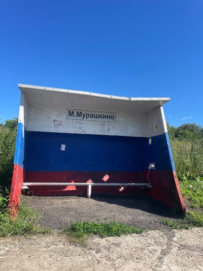 A bus stop structure, painted in the colors of the Russian flag (white, blue, and red), is situated outdoors under a clear blue sky. The structure has a white roof and a white upper back wall where a sign reads "М.Мурашкино" (M. Murashkino). Below the sign, the wall is painted blue, and the lower section is red. The structure has an open front and is weathered, with peeling paint and some graffiti visible. A white bench is attached to the back wall of the bus stop. The ground in front of the bus stop is covered with gravel, and grass and foliage grow around the structure and in the background. A paved surface is visible in the foreground. The scene depicts the bus stop in the settlement of Malyye Murashki, Russia.