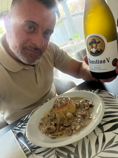 A man with short-cropped hair and a grey beard is holding a bottle of white wine, labeled "Faustino V Rioja." He is wearing a beige polo shirt and is seated at a table. In front of him is a white plate with food that appears to be a pastry topped with mushrooms in a creamy sauce. The setting seems to be outdoors on a patio or balcony with furniture and plants visible in the background. The lighting suggests it is daytime. The image does not contain enough visual information to determine the specific location within Junglinster, Luxembourg.