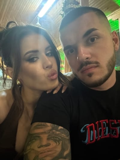 A close-up, waist-up selfie depicts a man and a woman in an indoor setting, identified as Debrashtitsa, Bulgaria. The woman, positioned on the left and slightly behind the man, has long dark hair styled in a partial updo with strands framing her face. She wears noticeable eye makeup, including winged eyeliner and defined eyebrows, and her lips are pursed in a "kissing" gesture with dark lipstick or gloss. A large, gold-toned hoop or disc earring is visible on her left ear. Her attire appears to be a dark brown or bronze halter-neck garment. Her right hand, with light-colored manicured nails, rests on the man's left shoulder.
The man, centrally positioned to the right of the woman and closer to the camera, has a close-cropped dark fade haircut and a neatly trimmed dark beard with a distinct 'soul patch' below his lower lip. He wears a black t-shirt with a partially visible embroidered logo on the chest in red and light blue, appearing to spell "DIESEL". An extensive, detailed sleeve tattoo, depicting stylized faces and figures, covers his left arm (visible on the right side of the image). He looks directly at the camera with a neutral expression.
The background features a wooden plank ceiling with a bright white fluorescent tube light fixture visible towards the top. Green accent lighting illuminates the upper right side of the frame, indicating artificial lighting. A textured wall, possibly brick or stone, is visible behind the woman. A decorative object, resembling a small, ornate crown or plant fixture, hangs from the ceiling near the top center. The overall scene suggests a casual social interaction, possibly in a restaurant or bar environment.