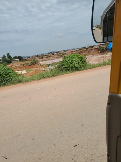 The scene depicts a landscape in Reifield-Ist, Nigeria, observed from the interior of a vehicle, likely a tricycle, positioned on a paved road. The perspective is angled slightly upwards and to the left.
In the foreground, a light grey-brown asphalt road occupies the bottom half of the image. On the far right, a vertical yellow structural element of the vehicle and a lower grey interior panel are visible. A black-framed rearview mirror is prominently positioned in the upper right, reflecting a road behind the vehicle and a distant white vehicle (possibly a bus or van) approaching.
The midground features green bushes and grass lining the left edge of the road, beyond which the terrain descends into an extensive area of reddish-brown, disturbed earth, indicative of excavation or significant erosion. Within this area, several irregularly shaped, reddish-brown water bodies are visible. Further in the mid-distance on the left, a few small, simple buildings, one with a light-colored roof, are situated on higher ground.
The background consists of a uniformly overcast, light grey sky. The distant horizon reveals faint outlines of rolling hills or elevated land, sparsely dotted with structures, suggesting a peri-urban or transitioning rural environment. No individuals are directly visible in the landscape. The overall environment suggests an area undergoing land development or impacted by natural processes in a tropical setting.