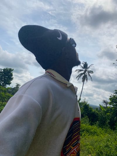 A person wearing a dark cap and a white polo shirt is looking up at a coconut palm tree. The background is a cloudy sky and lush green vegetation, consistent with the environment of Achara, Nigeria.
