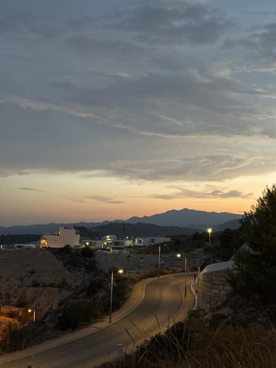 The image captures a dusky twilight scene in Finestrat, Spain, featuring a winding road leading through a hilly, residential area. The sky is a canvas of muted grays and soft oranges, with streaks of clouds suggesting the last light of day.

In the foreground, a paved road curves gently upwards, lined with streetlights casting warm pools of light onto the asphalt. The roadside vegetation is dry and sparse, with hints of taller grasses in the lower right corner. A low stone wall, constructed with rough-hewn blocks, borders the road on the right, forming a retaining wall.

Further up the hill, clusters of modern buildings are visible. One prominent white building stands out, its windows illuminated, suggesting habitation. Other structures, appearing to be houses or apartment blocks, are scattered across the hillside, their lights adding to the serene atmosphere. In the distance, a range of mountains forms a dark silhouette against the fading sky. The overall impression is one of a quiet, developing residential area on the outskirts of a Spanish town, bathed in the ambient light of dusk.