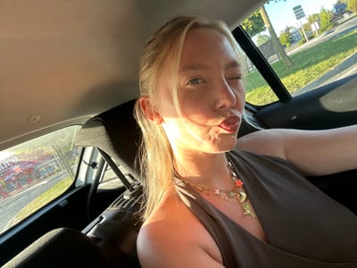 A close-up selfie captures a blonde woman winking and pouting her lips while seated in the back of a car. Her face is illuminated by warm, golden sunlight entering through the window, creating highlights on her hair and skin, and casting some shadows. She has light-colored eyes.
She is wearing a dark-colored, possibly brown or dark gray, halter-neck top. Around her neck are multiple layered gold necklaces adorned with various charms, including a red starfish, a seashell, a sun, a hummingbird, and small white flowers. Her blonde hair is styled in a ponytail or pulled back, with some strands framing her face.
The setting is the interior of a car, seen from the perspective of a passenger in the back seat. Parts of the car's headliner, a black car seat, and a seatbelt are visible. Through the car windows, an outdoor scene unfolds under a bright, sunny sky, indicating daytime, likely late afternoon given the quality of the light.
The view outside the window shows a road, green grassy areas, and trees. In the background, there's a road sign indicating directions to various locations, including "WASQUEHAL," "ANSTAING," "LEERS," and "ASCQ," confirming the location context of Leers, France. Another visible detail is a red building in the distant background, partially obscured, which appears to have "DRIVE" written on it, suggesting a drive-through establishment. A car window sticker with "E3 43R-00029" is also partially visible. The overall mood of the image is playful and bright.