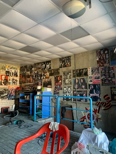The image depicts a cluttered interior space, possibly a garage or workshop, with walls covered in numerous rock and roll band posters. The ceiling is a suspended tile system. In the foreground, a red plastic chair is prominently displayed, with a pair of white work gloves draped over its back. To the left of the chair, on the floor, are various tools and equipment, including a red radio, a black vacuum cleaner, and a green and black hammer drill. A blue shelving unit stands against the back wall, filled with audio equipment and books. The walls themselves are adorned with a dense collection of posters, featuring bands like KISS, Led Zeppelin, Deep Purple, Queen, and Metallica. The floor is paved with interlocking grey bricks. In the background, a large medal with orange and black ribbon is attached to the wall, suggesting a patriotic or commemorative context. The scene appears to be set in a residential or semi-industrial area in Afanasyevo, Russia, given the presence of Russian text on some posters and the medal.