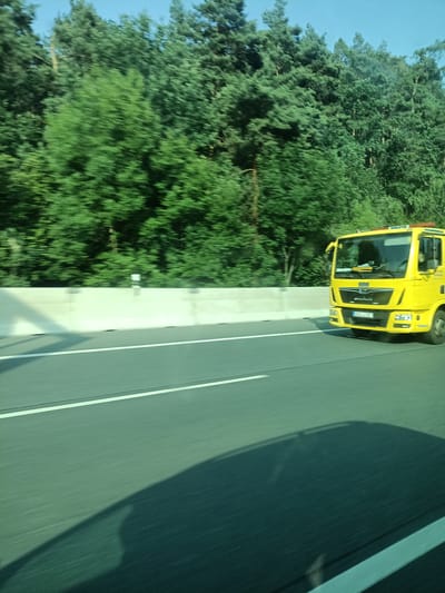 The image shows a yellow tow truck driving on a highway through a wooded area. The tow truck is the main subject of the image, and it is captured in motion, indicated by the motion blur. The scene is set on a sunny day, as suggested by the bright lighting and the clear blue sky visible in the background. The highway has clear white lane markings, and a concrete barrier separates the road from the trees. The trees are lush and green, typical of a temperate climate during the summer months.
There is some text visible on the tow truck's license plate, which appears to be "BGL L 107". There are also some white signs on poles along the side of the highway, but the text on them is not legible due to the distance and motion blur. The overall mood of the image is neutral, depicting a common scene of transportation on a highway. The time of day appears to be daytime, possibly mid-morning or afternoon, given the quality of the light. The location context is Kunice, Czechia.