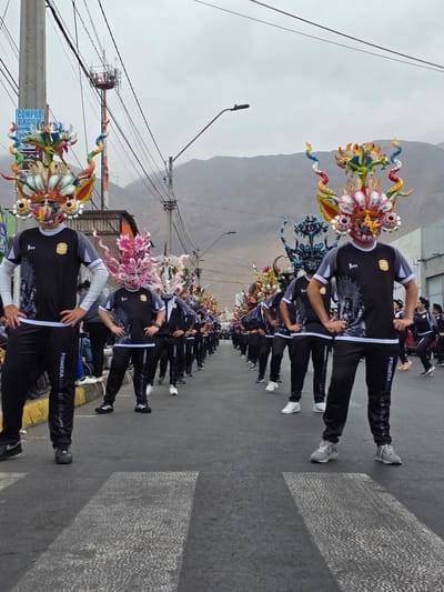 This image captures a vibrant street parade, likely in Iquique, Chile, given the traditional masks and attire. In the foreground, two individuals, predominantly men, are positioned with their hands on their hips, facing the camera. They are dressed in matching black tracksuits with grey trim and white accents. Their tracksuits feature a distinctive emblem and text that reads "PRIMERA DIA" and "ALANZA," suggestive of a team or organization. Both wear large, elaborate, and colorful masks that cover their faces. These masks are fantastical, resembling mythical creatures with multiple eyes, horns, and brightly colored, flowing headdresses that extend upwards and outwards.

Behind these two individuals, a procession of similarly attired participants, also wearing elaborate masks, stretches down the asphalt street. The masks vary in design and color, adding to the visual spectacle. The street itself is marked with white pedestrian crossings. The surrounding environment suggests an urban setting with buildings on the right side of the street, some appearing commercial or residential. Power lines crisscross the sky, and in the background, a mountainous, arid landscape is visible under an overcast sky. A tall communication tower with antennae is also present in the background, near a utility pole with a "COMPRO VEHICULOS" (Buy Vehicles) sign. Spectators are sparsely visible on the left side of the street, some seated on a yellow curb. The overall atmosphere is one of a festive cultural event.