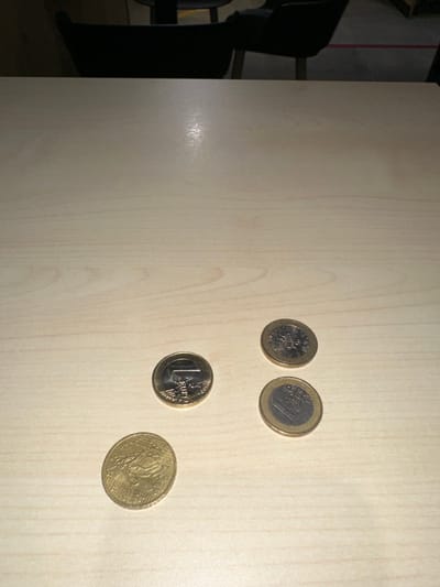 Four euro coins are scattered on a light-colored, possibly wooden surface. They include a 10-cent coin, a 20-cent coin, a 50-cent coin, and a 1-euro coin. The coins appear randomly placed, with slight overlaps between them. The background shows a blurred room setting with a chair visible on the left edge. The lighting highlights the coins and part of the surface.
