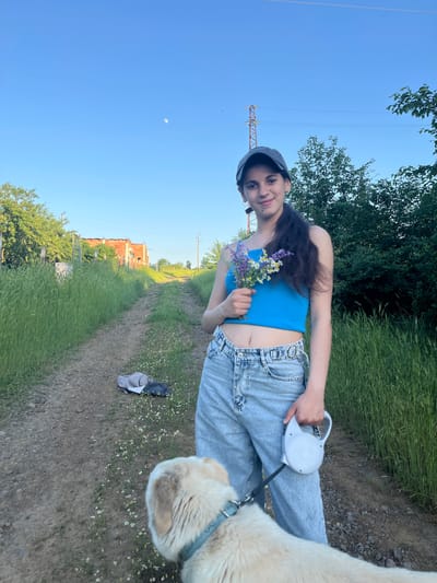 The photo shows a young woman in Lozarevo, Bulgaria, standing on a dirt path bordered by tall grass. She is wearing a blue tank top, light blue jeans, and a gray cap. She holds a small bouquet of wildflowers in one hand and a retractable dog leash in the other. A cream-colored dog is partially visible to her right. In the background, there is a rural landscape with trees, a blue sky, and a building with an unfinished brick facade. Power lines and a transmission tower are visible on the left side of the image. A discarded pile of fabric lies on the path near the dog.