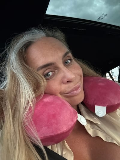 A selfie is taken inside a car, looking up towards the sunroof. A woman with blonde hair, partially graying at the temples, is looking directly at the camera. She is smiling and resting her head against two pink neck pillows. She is wearing a light-colored, collared shirt. The interior of the car is dark, and the sky is visible through the sunroof, with some clouds. There is a sticker on the sunroof with text and a QR code. The background outside the car is blurry but appears to be a street scene. The image does not provide any specific visual cues that would allow for an accurate determination of the location within Ban Nong Mu, Thailand.