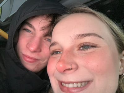 Two individuals are closely positioned with their faces near each other. The person on the left has blonde hair, fair skin with freckles, blue eyes, and a nose ring. They are smiling, revealing their teeth. The person on the right is wearing a dark hoodie, and their face is partially obscured by the hood. They also have fair skin and blue eyes. The image appears to be rotated slightly, and the composition focuses on the faces of the two people.