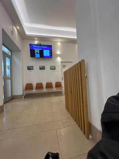 A waiting room features four tan chairs aligned against a white wall decorated with framed images. Above the seating, a light wood slat screen hangs from the ceiling. A television displaying text in a foreign language is mounted on the wall. The room is bright with a tiled floor. A window or glass door sits at the bottom of the image.