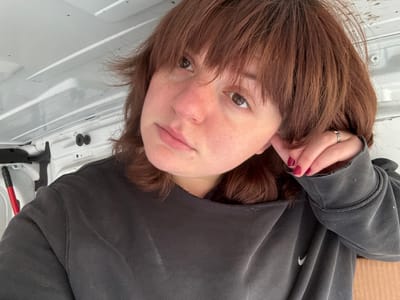 A single individual, appearing female, is depicted from the chest up within the white, utilitarian interior of a vehicle, likely a van or cargo space. The individual has reddish-brown, medium-length hair with bangs and fair skin, showing some freckles. A silver-colored nose ring is visible on her left nostril. She wears a faded dark gray crew-neck sweatshirt, exhibiting wear along the collar and cuffs, with a white Nike swoosh logo visible on the lower right front. Her left hand is raised, with fingers gently touching her hair near her left ear; the nails are painted dark red, and a silver-colored ring with an intricate design is present on her left ring finger. The individual's gaze is directed upwards and to the right of the frame, with a neutral to contemplative expression. The background consists of white paneled walls and a ceiling with visible structural contours. A red-handled tool is partially visible in the lower-left corner of the frame, leaning against the wall. The image provides no specific visual information to confirm the location is Cortona, Italy.