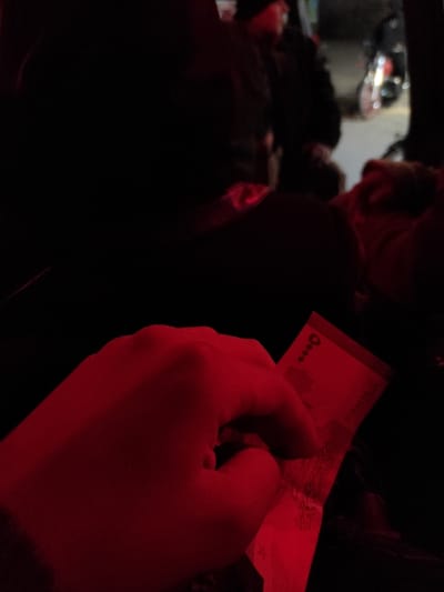 A hand holds a currency note, illuminated by a red light. The note's details are partially visible, showing a number "5000." The background is dark and blurred, with glimpses of a blurry environment. The red lighting casts a strong hue over the scene, creating a stark contrast with the dark backdrop.