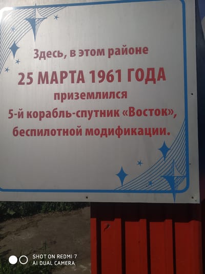 The image shows a sign that reads "Здесь, в этом районе, 25 МАРТА 1961 ГОДА приземлился 5-й корабль-спутник «Восток», беспилотной модификации." The sign is white with red text and blue accents. It's attached to a red structure with vertical slats. In the foreground, there's also some vegetation and asphalt. The sign commemorates the landing of the Vostok 5 spacecraft, an unmanned version, in the area on March 25, 1961.