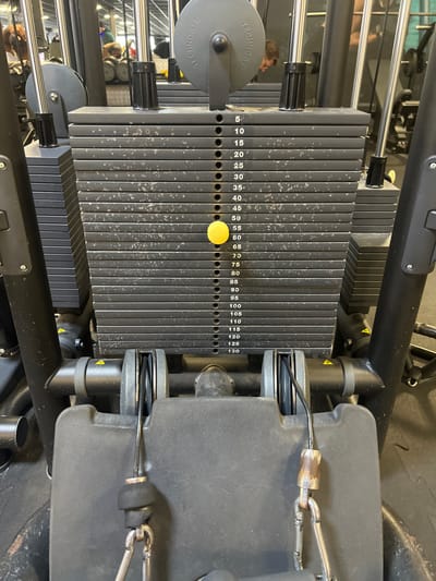 The image depicts a close-up, eye-level view of a weight stack on a Technogym cable fitness machine located in a gym in Wolfisheim, France. The weight stack consists of approximately 26 dark gray, rectangular metal plates, each marked with a number in increments of 5, ranging from "5" at the top to "130" at the bottom. A bright yellow selector pin is inserted into the hole corresponding to the "55" mark. The plates exhibit signs of wear, including scuffs, scratches, and light dust accumulation. Above the weight stack, a black Technogym branded pulley wheel is visible, guiding a cable. Below the weight stack, the base of the machine shows two smaller pulleys with cables leading to two metallic carabiner clips, which are attached to worn, black rubberized handles or straps. The foreground is occupied by the machine's black frame and padded elements. In the blurred background, reflections on metallic surfaces indicate other gym equipment, including dumbbells, and indistinct figures of individuals, suggesting an active gym environment with artificial lighting. No direct human interaction with the depicted machine is occurring in the foreground.