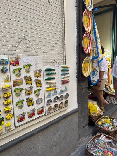 In Pogerola, Italy, a store display features various souvenir items, primarily magnets and decorative plates. The display includes a white grid backing from which items are hung or attached. On the left, multiple rows of magnets are visible, depicting scenes of coastal towns, vehicles, and lemons. Prices are indicated with handwritten tags. To the right, decorative ceramic plates with lemon motifs are hanging, accompanied by a fabric item featuring a similar design. Further to the right, a woman in a white top and tan skirt walks past the display. Baskets filled with colorful balls, lemons, and wrapped goods are situated on the ground near the store entrance. The scene suggests a tourist-oriented retail environment.