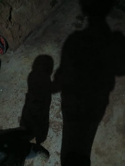 This image, captured in Witeithie ward, Kenya, depicts a dimly lit scene dominated by prominent shadows on a textured ground surface. The elongated shadows of an adult figure stand vertically on the right, with a smaller child's shadow positioned to its left, appearing to be in close proximity or interaction. In the lower left quadrant, a dark-bodied turtle or tortoise is partially visible, displaying a textured, possibly patterned, dark green and brown head with a discernible eye and snout. A red and black sandal is visible in the upper left corner, partially obscured. The ground surface is a rough, uneven concrete or similar hard material, showing variations in tone and texture. The upper left background features a dark, uneven wall or earth bank.