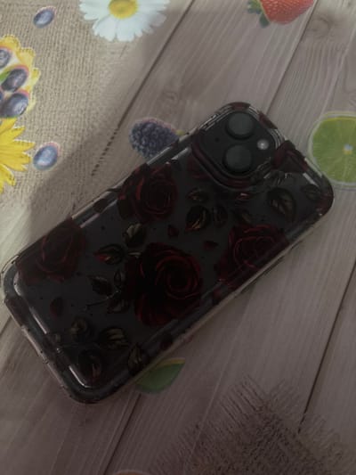 The scene, located in Ihtiman, Bulgaria, depicts a smartphone encased in a clear protective cover, resting on a decorative surface. The smartphone, observed from its rear, features a dual-lens camera system and a flash module in the upper section. The clear case encloses a dark-colored phone, with the case itself having a printed design of deep red roses, accompanying green leaves, and scattered dark red petal-like elements on a dark grey or black background.

The decorative surface beneath the phone is composed of two primary sections. The left portion exhibits a textured, light beige or tan fabric-like material, possibly a placemat, adorned with printed imagery including a white daisy with a yellow center, several individual blueberries, and a cluster of blueberries within a yellow, possibly floral, outline. The right portion of the surface displays a lighter, wood-grain pattern with diagonal lines, suggestive of a wooden table or a wood-patterned mat. This section features additional printed imagery, including a red strawberry, a green lime slice, and a blackberry.

The overall lighting is dim, casting soft shadows and contributing to a subdued visual tone. No human subjects or explicit actions are present within the frame.