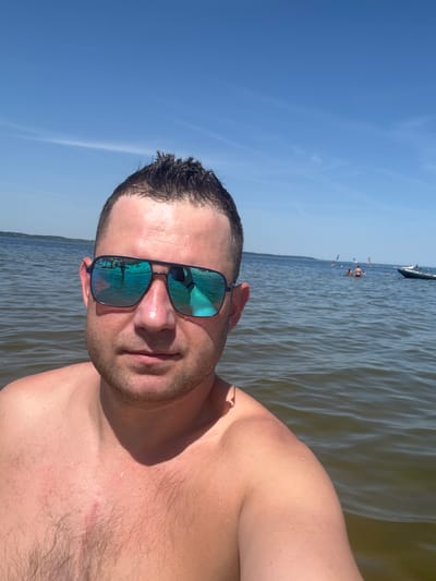 A man is taking a selfie in the water near the beach in Rewa, Poland. He is wearing mirrored sunglasses and is shirtless. The water is calm, and the sky is clear and blue. In the background, several people are visible in the water, some engaging in water sports like windsurfing. A boat is also present in the distance. The scene appears to be a sunny day at the beach.
