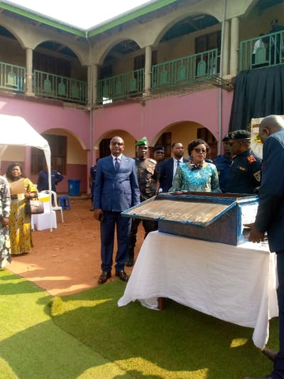 The photo shows a group of people standing in a courtyard.  The people are standing in front of a table draped in a white tablecloth. On the table is a blue metal box with writing on it. The people are all wearing formal clothing.  Some have military attire.  The background shows a pink building with arched doorways and green balconies.  The ground is dirt, and there is artificial turf at the bottom of the photo.  The photo appears to have been taken in Beni, Democratic Republic of Congo.  The coordinates provided in the image description are: 0.491, 29.483.