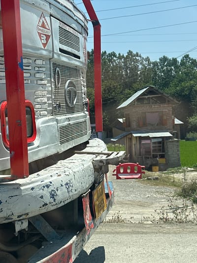 In the city of Khrewan Lasi Pora, India, a weathered, white truck is parked on a dusty road. The truck's front is visible from the side, with red metal supports framing its exterior. A diamond-shaped hazard symbol indicating "FLAMMABLE LIQUID" with a "3" inside is prominently displayed on the truck's body, along with additional warnings that are partially obscured.
The truck appears aged, with chipped paint and signs of wear. Behind the truck, a simple, two-story house made of brick and corrugated metal roofing stands against a backdrop of lush green trees and a bright blue sky. A small, red, child-sized bench sits on the side of the road near the house. The ground is a mix of gravel and dirt, with some dry vegetation and scattered small pink flowers. The overall scene suggests a rural or semi-rural setting under a clear, sunny sky.