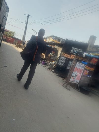 A man wearing dark clothes and carrying a bag walks on a paved street. Behind him are buildings and structures, including a sign with text. Overhead are utility poles and wires against a light sky. The perspective is angled, giving a tilted view of the scene. The image appears to be taken outdoors during the day.