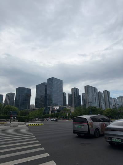 The image shows a street scene in Binjiang District, China. The sky is overcast with heavy clouds. High-rise buildings line the street. Cars are parked on the right side of the road, and a crosswalk is visible. A car decorated with Hello Kitty stickers is among the vehicles. Pedestrians can be seen walking on the sidewalk.