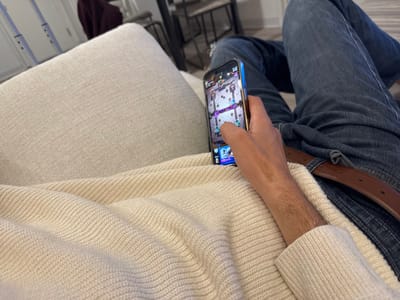 An individual is depicted from a first-person perspective, reclining in an indoor setting. The subject's right arm, partially covered by a light cream-colored ribbed knit sweater, holds a smartphone. The phone, identifiable as an iPhone due to its design and a distinct blue bezel, displays the active user interface of the mobile strategy game "Clash Royale." The individual's right thumb is positioned on the screen, indicating interaction with the game. Below the phone, a brown leather belt is visible over blue denim jeans, which are worn by the subject. The person is resting on a light-colored, textured fabric cushion or sofa, which forms the foreground and left side of the image. The background is softly out of focus, showing indistinct elements of a domestic interior, including what appears to be a dark garment draped over furniture and light-colored walls. The specific location within the city of Washington, United States, cannot be determined from the visual evidence in the image.