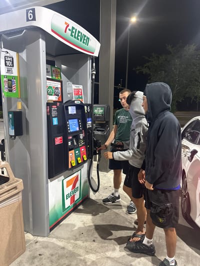 The image shows a scene at a 7-Eleven gas station, with a gas pump displaying $15.00 and 15.247 gallons pumped. Two young males stand near the pump, while another person, possibly filling a white car, is seen. The gas station offers various promotions, including savings on Monster Energy drinks and nicotine products, as well as extra discounts for using the 7-Eleven app. Hand sanitizer is available at the pump. The pump is labeled with the number "6." Based on the "USF Numa" text on a shirt, it is unlikely that the image is in the city of Homosassa Springs, United States of America.
