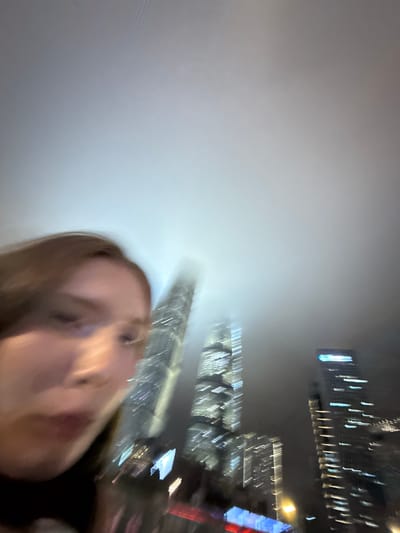 This is a blurry, low-light, vertical-orientation photograph taken at night in Pudong, China. The main subject is the face of a young woman, who is looking towards the camera with her mouth slightly open. Her facial features are indistinct due to the blur. She appears to be outdoors, with a cityscape in the background.
The background consists of a dense fog or mist that obscures most of the scene, creating a bright, diffused glow in the upper left quadrant of the image. Emerging from the fog are the illuminated upper floors of skyscrapers, their lights appearing as streaks and blurs. The buildings suggest a modern urban environment. One building in the distance has prominent blue and white lights. Another building shows a pattern of white lights. A faint yellow light from a streetlamp is also visible. The overall impression of the background is that of a city at night, possibly shrouded in fog or smog.
The motion blur suggests that either the camera or the subjects were moving during the exposure, contributing to the abstract and ethereal quality of the image. The time of day is clearly night, and the weather appears to be foggy or misty, which is common in large urban areas. The location is identified as Pudong, China, known for its distinctive skyline. There is no visible text in the image. The mood is somewhat mysterious due to the blur and fog, but the woman's direct gaze might imply a sense of engagement or surprise.