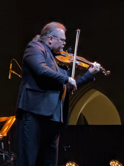 A man in a dark suit plays a violin on stage. He has glasses and light hair pulled back in a ponytail. The violin and bow are prominent. A microphone stand is visible near the bottom of the frame. The background is a mix of light and dark areas, including a curved light fixture and stage lighting. The image is slightly rotated counter-clockwise.