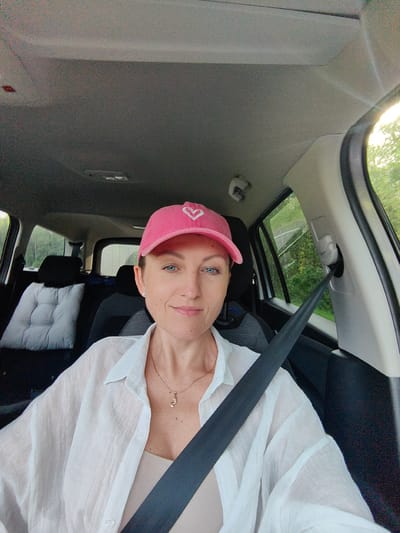 A woman is sitting in the driver's seat of a car. She is wearing a pink baseball cap with a white heart on it, a white semi-sheer button-down shirt, and a beige tank top. She has on a gold necklace with a small cat pendant and small diamond earrings. The car interior is visible, with a grey headliner, black seats, and a seatbelt across her chest. Outside the car windows, trees and greenery are visible, suggesting a rural or suburban setting. The lighting indicates it may be late afternoon or early evening. The specific location of Хаценчыцкі сельскі Савет, Belarus, is not ascertainable from the visual information provided in the image.