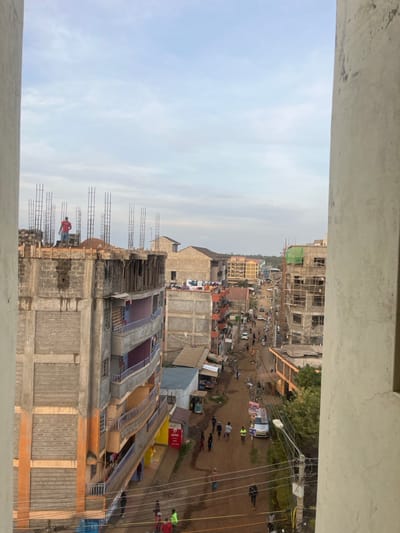 An elevated, downward-angled view captures an urban scene within Gitothua ward, Kenya, characterized by mixed-use development and ongoing construction under an overcast sky. In the immediate foreground, a concrete structural element frames the left side, with a similar element partially visible on the right. Directly below and to the left, a multi-story building, grey and unpainted, is actively under construction, showing exposed concrete blocks and numerous vertical rebar elements on its uppermost level. A lone male figure, dressed in a red shirt, stands on this building's rooftop amidst construction materials.
The central and midground reveal a bustling unpaved dirt road winding into the distance, bordered by a dense array of multi-story structures. Many of these buildings are in various stages of completion, displaying exposed concrete and brickwork, some with visible rebar and scaffolding, including a structure on the right with green netting. Numerous pedestrians, including adults and children, traverse the dirt road. Several vehicles, primarily white cars, are also present on the thoroughfare. Utility poles with a network of overhead wires line the street. Small commercial kiosks and storefronts, one prominently displaying an "Airtel" sign, are situated at street level. Clothes are visible drying on balconies of some residential-style buildings. The background features a continuation of densely packed buildings, fading into the hazy distance beneath a uniformly cloudy sky.