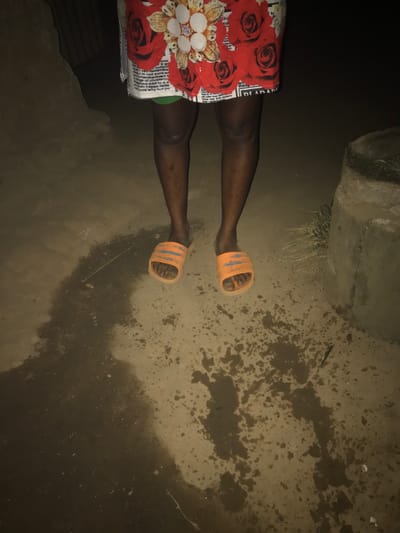 A person in orange sandals stands on a wet, muddy surface. A concrete or stone structure is in the upper left corner. The person wears a skirt or wrap with a red floral pattern and newspaper print. The surrounding area is dark, with the ground appearing muddy and uneven. The picture is taken from an overhead perspective.