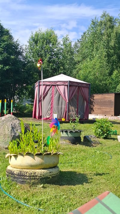 The image shows a backyard scene in Novovilgovskoye Sel'skoye Poseleniye, Russia, on a sunny day. In the foreground, there is a large rock to the left and a planter made from two stacked tires filled with green plants and a small solar-powered light. A colorful pinwheel is planted in the ground near the planter.
In the midground, a hexagonal gazebo with a light-colored roof and maroon curtains is set up on a wooden deck. To the right of the gazebo, there is a wooden shed. A second planter, also made from tires, sits to the right of the gazebo. Green grass covers the ground, with scattered rocks and bushes throughout the yard.
In the background, dense green trees form a natural border around the property. The sky is blue with scattered white clouds. Several colorful poles are visible behind the large rock in the foreground. No people are present in the image.