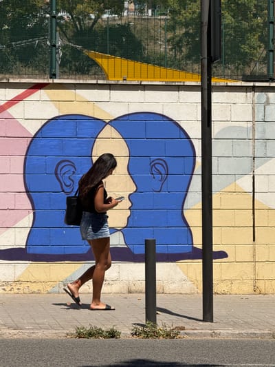 A woman walks along a sidewalk in front of a mural on a brick wall in l'Hospitalet de Llobregat, Spain. The mural features two large, blue, profile silhouettes of human heads facing each other. The woman, wearing a black sleeveless shirt, denim shorts, and black sandals, is looking down at her mobile phone as she walks from left to right. She carries a black bag over her left shoulder. The sidewalk is made of concrete pavers, and a street is visible in the foreground. Behind the mural, a metal fence encloses an area with green trees and some yellow structures. Two black metal poles stand on the sidewalk, one partially obscuring the mural. The wall itself is painted with geometric patterns in pink, yellow, and white bricks, in addition to the blue heads. The lighting suggests it is daytime with sunlight casting shadows.