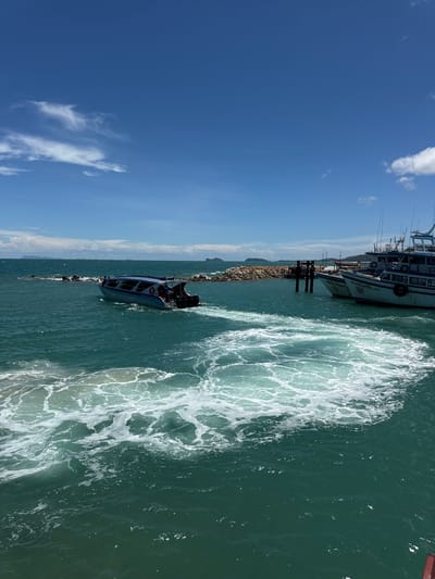 A speedboat is maneuvering through turquoise water, creating a foamy wake. In the background, two passenger boats are docked near a stone jetty. The sky above is a clear blue with scattered white clouds. The location is Ban Haad Rin, Thailand.